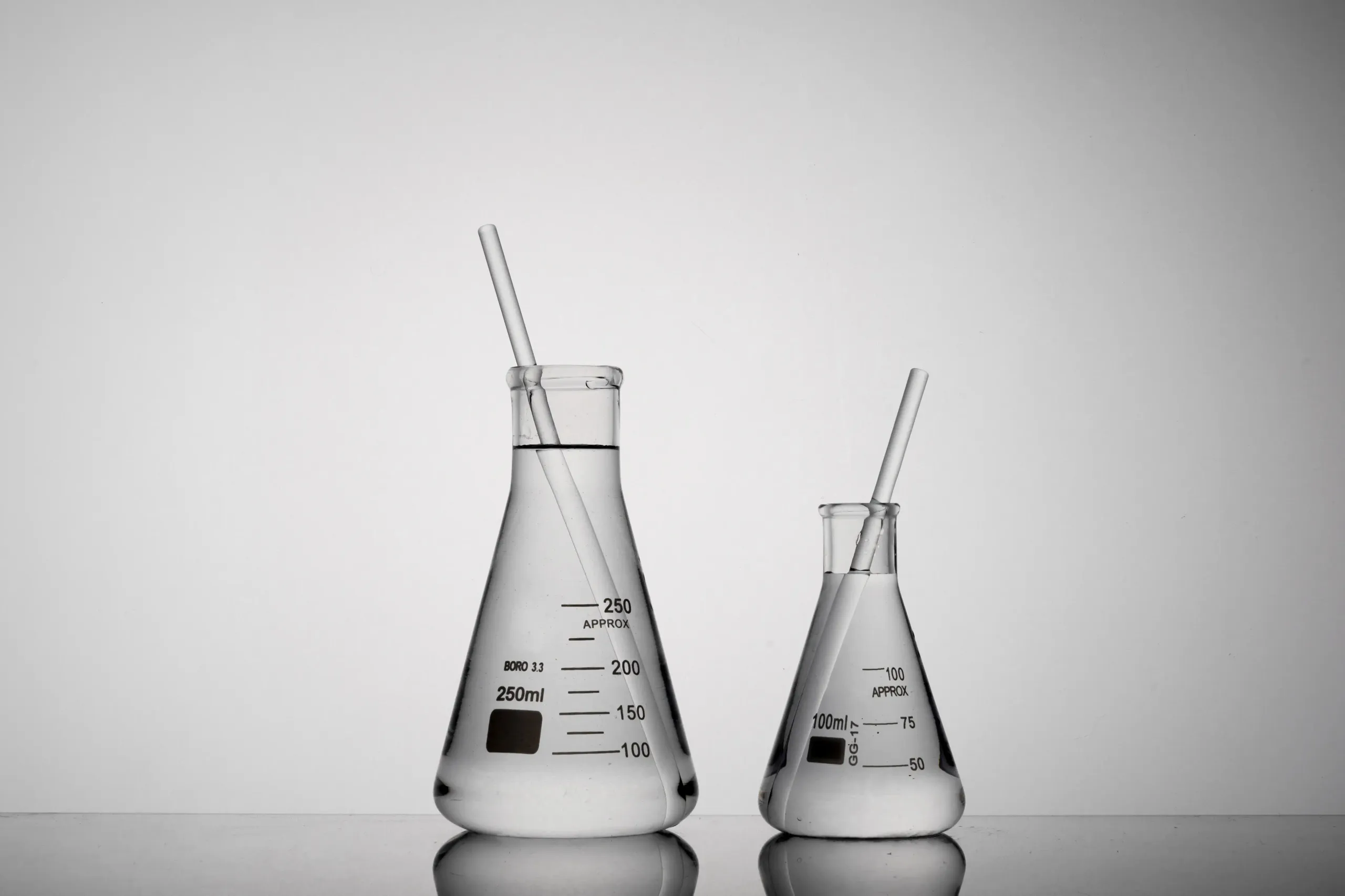 Zwei Labor-Erlenmeyerkolben (250 ml und 100 ml) mit klarer Flüssigkeit und Glasrührstäben auf neutralem Hintergrund.